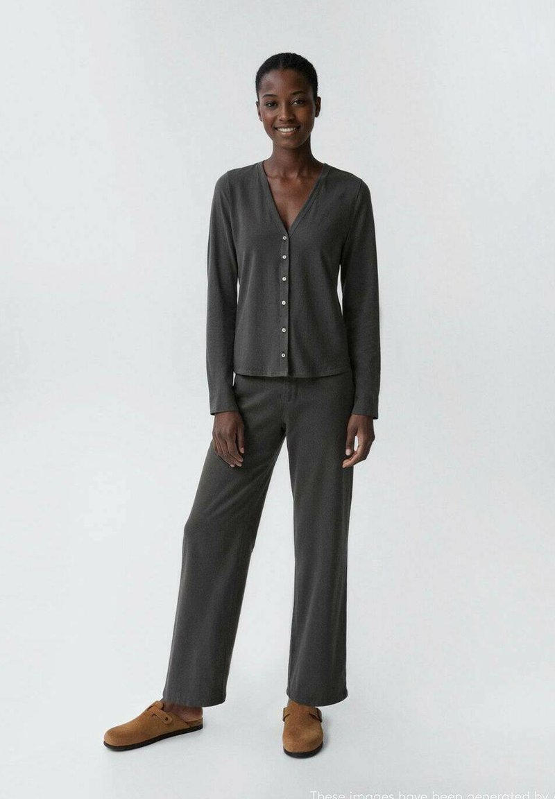 Smiling woman stands wearing dark gray buttoned long-sleeve top, matching wide-leg pants, and brown slip-on shoes on white background.