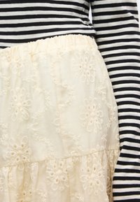 Κοντινό πλάνο κρεμ φούστας με κεντήματα λουλουδιών, φορεμένη με μακρυμάνικο τοπ σε ασπρόμαυρες ρίγες.