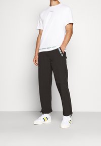 Vitt bomullströja med grön logotyp, kombinerat med svarta byxor med gröna och vita detaljer, samt vita sneakers med gröna och gula ränder.