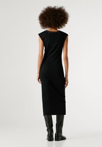 Abito midi nero aderente con maniche a cappuccio, caratterizzato da borchie decorative attorno al collo. La modella indossa stivali in pelle nera fino al ginocchio.