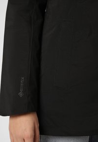Schwarze wasserdichte Jacke mit strukturiertem Finish, ausgestattet mit einer Tasche und einem sichtbaren Gore-Tex-Logo am Ärmel. Taillierte Passform.