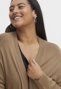 Hnedý, oversized cardigan s rebrovanou textúrou a širokými klopami. Čierny vnútorný top viditeľný. Modelka má na sebe zlaté kruhové náušnice.