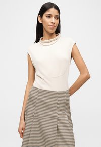 Γυναίκα που φοράει αμάνικο εκρού τοπ με μαργαριταρένιο κολιέ και ψηλόμεση φούστα με σχέδιο houndstooth, ποζάροντας μπροστά σε απλό ανοιχτό φόντο.