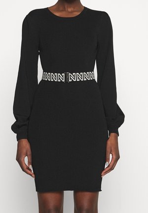 Czarna prążkowana sukienka z długim rękawem z bufkami oraz białym paskiem w talii z czarnym, powtarzającym się wzorem litery "N".