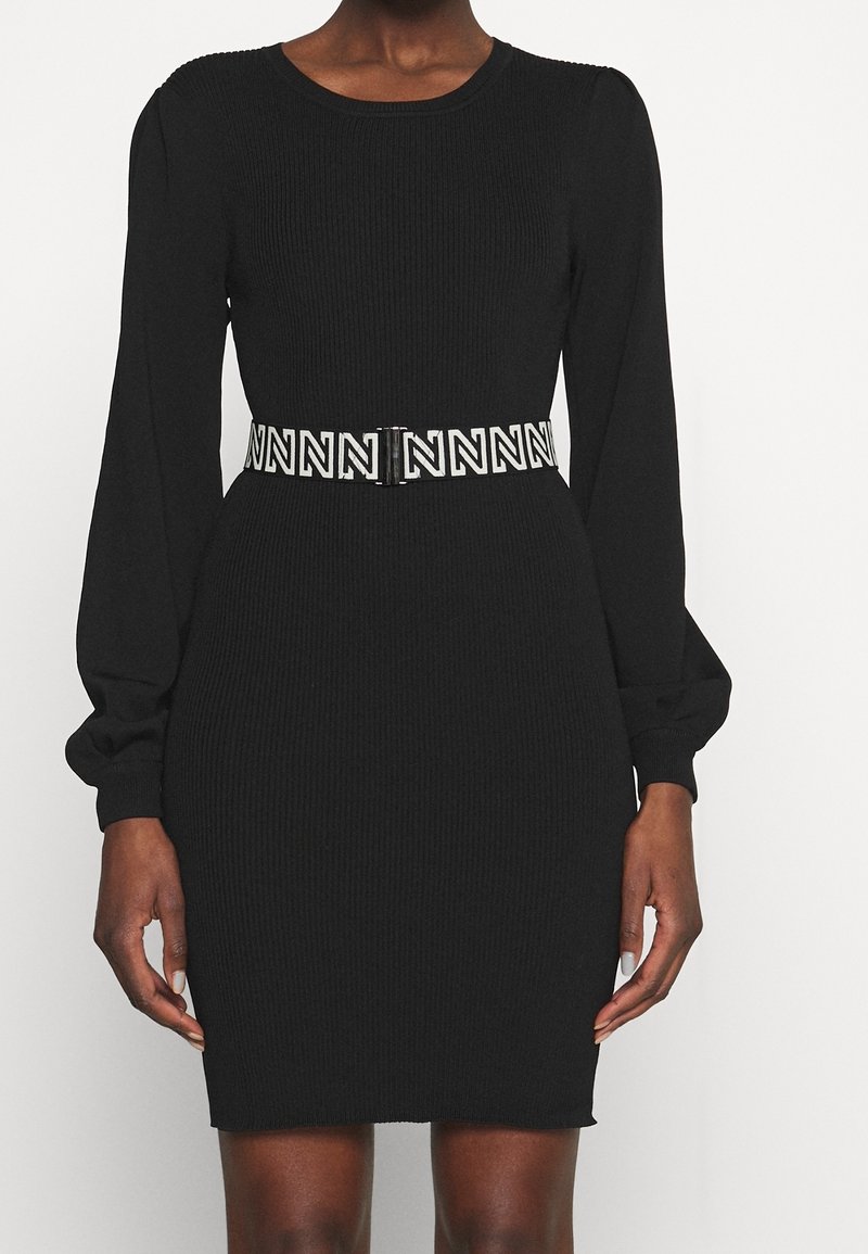 Robe noire à manches longues côtelées avec manches bouffantes et ceinture blanche ornée de motifs répétés en "N" noirs à la taille.