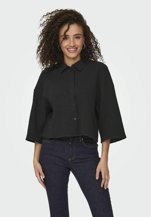 Black, cropped button-up shirt in textured fabric with a collar and three-quarter sleeves, paired with dark blue jeans.