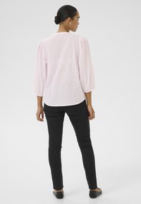 Blouse texturée rose clair avec des manches bouffantes, vue de dos. Associée à un pantalon noir ajusté et des chaussures plates noires. Design simple.
