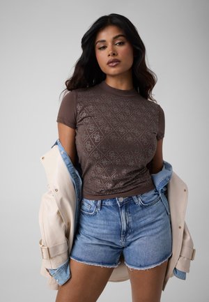 Femme aux longs cheveux foncés portant un t-shirt marron à motifs, un short en jean clair et une veste beige drapée sur ses épaules, sur un fond gris.