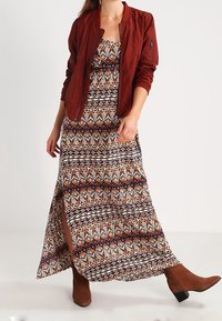 Bordeauxfarbene Bomberjacke über einem gemusterten Maxikleid mit geometrischen Designs in Braun, Orange und Marineblau, mit einem seitlichen Schlitz und braunen Stiefeln.