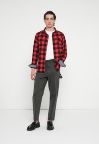 Rot-schwarz kariertes Flanellhemd, übereinander getragen über einem weißen T-Shirt, kombiniert mit grauen, schmal zulaufenden Hosen und schwarzen Schuhen.