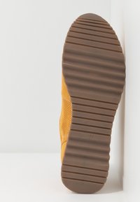 Zapatilla amarilla con un upper texturizado y perforaciones, suela de goma marrón con ranuras horizontales y un borde acolchado para mayor comodidad.