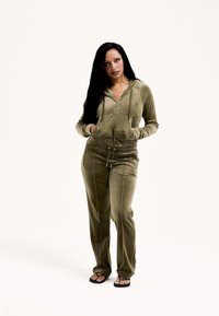 Zielono-oliwkowy dres z weluru, z bluzą na zamek z kieszeniami z przodu i sznurkiem w talii, zestawiony z czarnymi sandałami.