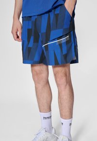 Blåmönstrade sportshorts med svart design, med en vit accentrand och en synlig logotyp på sidan. Bärs med vita strumpor och skor.