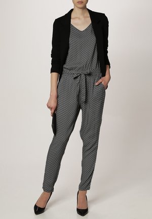 Woman wearing patterned black and white jumpsuit with a tied waist, black blazer, black heels, and holding a black clutch.