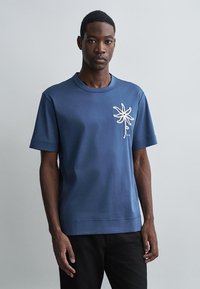 Camiseta azul de manga curta feita de algodão com decote redondo. Apresenta um bordado floral texturizado em branco no lado esquerdo do peito.
