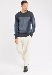 Maglione blu navy a maglia con strisce orizzontali blu, scollatura rotonda e tre bottoni decorativi sulla spalla, abbinato a pantaloni di colore crema.