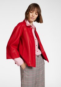 Giacca di pelle rossa con una vestibilità rilassata, dotata di colletto e chiusura con bottoni. Indossata sopra una camicia rosa chiaro e pantaloni a scacchi.