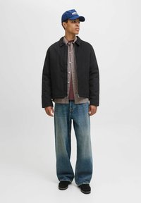 Giacca nera tagliata sopra una camicia a quadri, abbinata a jeans larghi blu. Un cappellino da baseball blu e scarpe scure completano l'outfit.