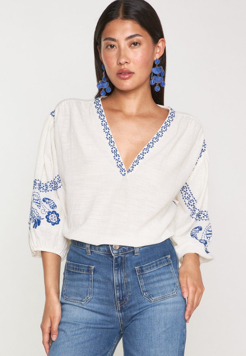 Blusa bianca in lino con ricami floreali blu sulle maniche e sul collo. Presenta un profondo scollo a V e maniche leggermente a sbuffo. Abbinate a un denim.