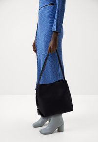 Svart tote-väska med slät textur och axelremmar, hålls av en person som bär en blå ribbad klänning och grå klackstövlar.