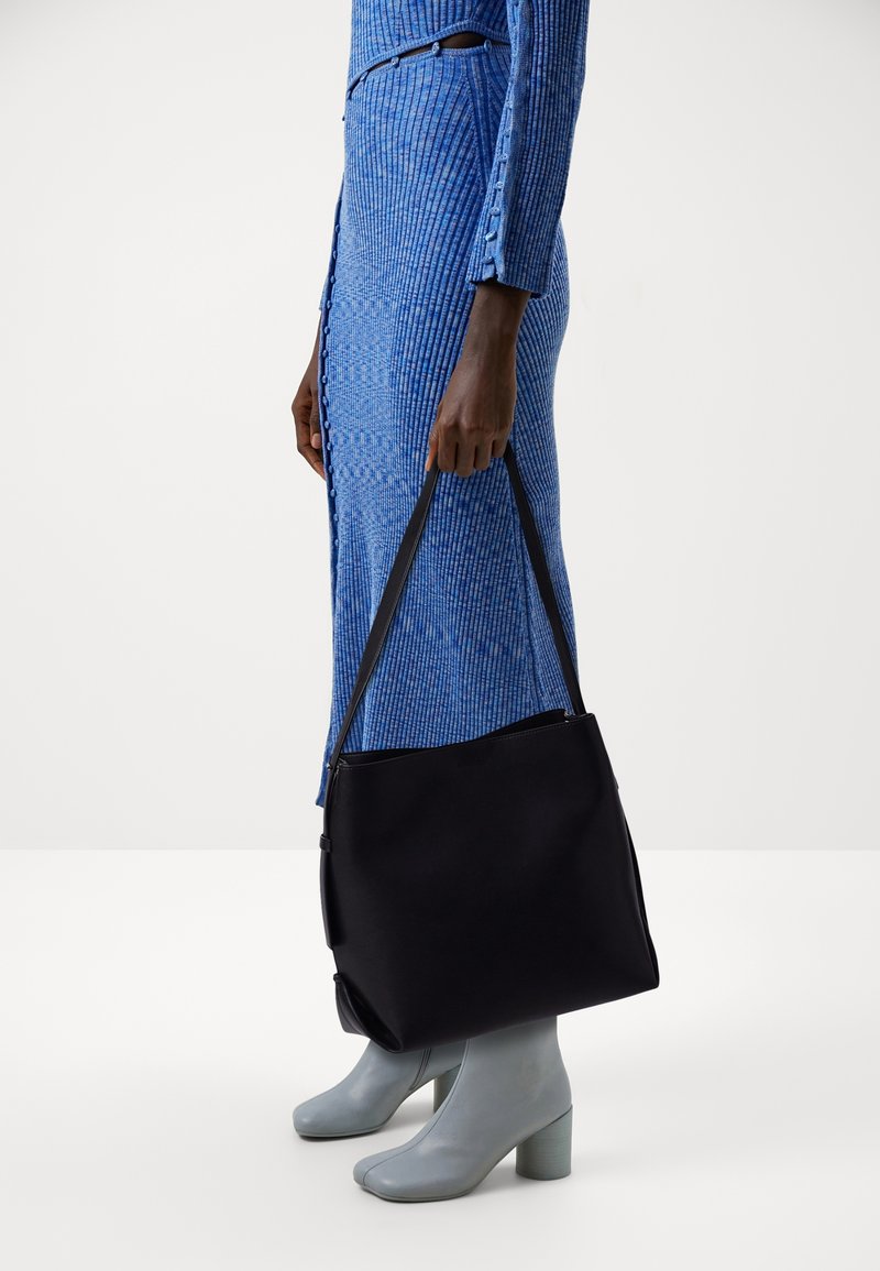 Svart tote-väska med slät textur och axelremmar, hålls av en person som bär en blå ribbad klänning och grå klackstövlar.