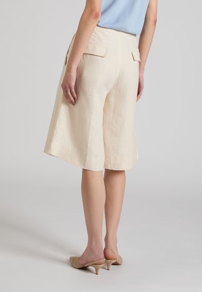 Mujer con pantalones cortos anchos beige hasta la rodilla con bolsillos traseros con solapa y zapatos slingback de tacón bajo beige, de pie contra un fondo liso.
