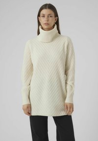 Valkjas turtleneck särk, millel on tekstureeritud chevron muster, pikad varrukad ja soonikuppud. Näidatud koos mustade pükstega.