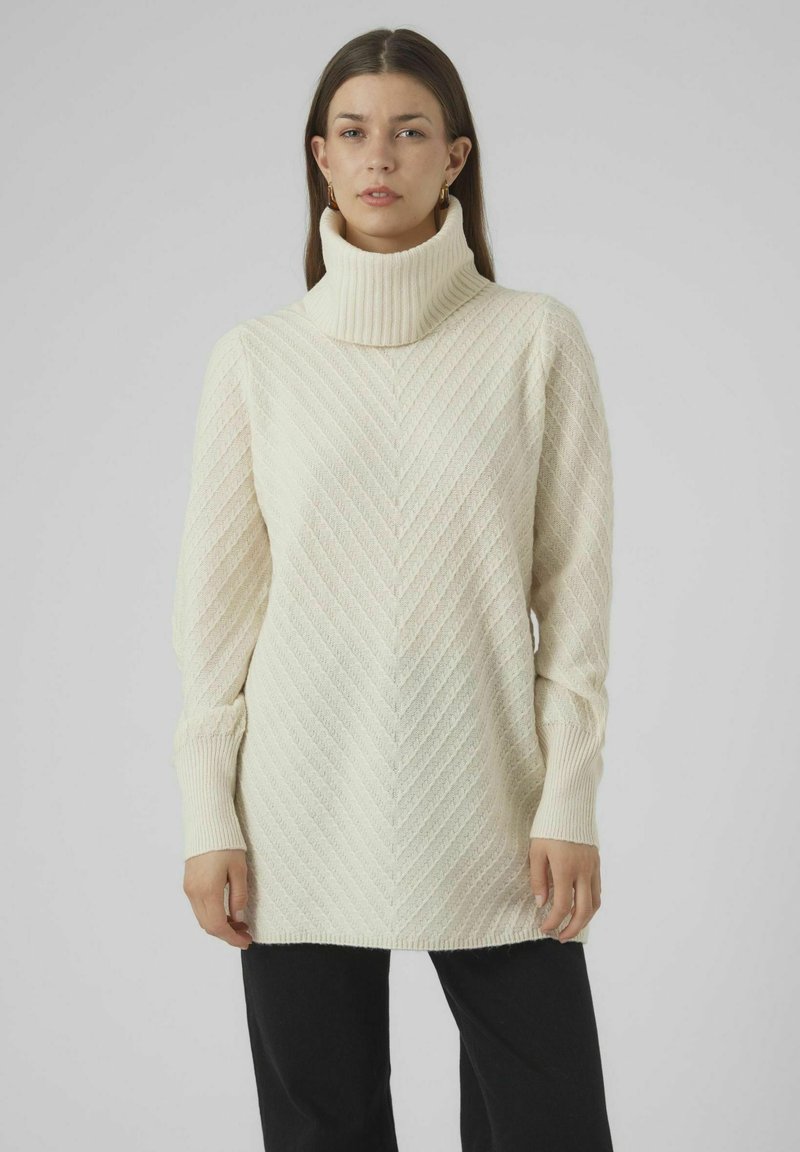 Valkjas turtleneck särk, millel on tekstureeritud chevron muster, pikad varrukad ja soonikuppud. Näidatud koos mustade pükstega.