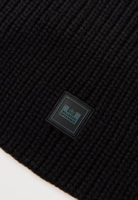 Czarna dzianinowa czapka z prążkowanym wzorem, z kwadratową naszywką w kolorze czarnym i turkusowym, na której widnieje napis „WEEKEND OFFENDER.”