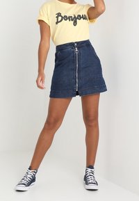 Gonna mini in denim con zip frontale e chiusura con bottone, abbinata a una maglietta gialla con testo in paillettes nere, insieme a sneakers blu.