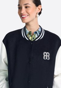 Fekete-fehér egyetemi stílusú dzseki bordázott gallérral, gombos záródással és hímzett "RB" logóval a mellrészen; virágmintás ing fölött viselve.