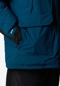 Teal isolerad jacka har en dragkedjeficka, 600 isoleringsmärke och svarta accenter. Materialet verkar slätt med en matt finish.