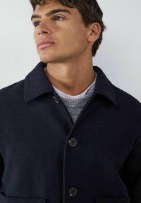 Granatowy wełniany płaszcz z kołnierzem, z zapięciem na guziki z przodu, dwiema bocznymi kieszeniami i teksturowanym szarym swetrem pod spodem.