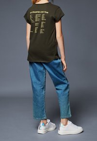 Tmavě zelené bavlněné tričko s krátkým rukávem, na zadní straně s žlutým textem uvádějícím města z turné. V kombinaci se světle modrými džíny a bílými teniskami s duhovými akcenty.