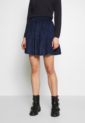 Kvinde iført en mørkeblå, rynket nederdel, sort langærmet top og sorte ankelstøvler med spænder, stående foran en hvid baggrund.