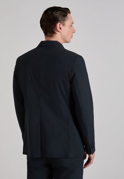 Man die zijwaarts staat en een nauwsluitend donkerblauw colbert en bijpassende broek draagt tegen een effen lichtgrijze achtergrond.