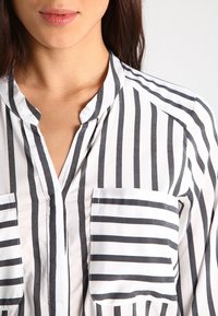 Camicia bianca con righe nere verticali, dotata di scollatura a V e due tasche frontali, realizzata in tessuto leggero.