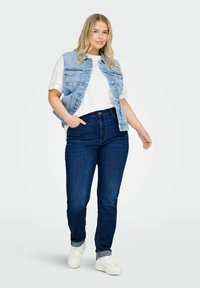 Moteris su ilgais šviesiais plaukais, vilkinti baltą marškinėlį, šviesiai mėlyną džinsinį švarką, tamsiai mėlynus džinsus ir baltus sportbačius, stovi su viena ranka kišenėje.