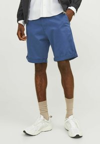 Shorts en coton bleu avec des ourlets roulés, associés à des chaussettes beiges et des baskets blanches. La tenue présente un design décontracté et des détails simples.