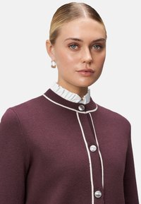 Burgundy cardigan with white trim and round buttons, featuring a textured fabric and high ruffled collar. Close-up view of the garment.
