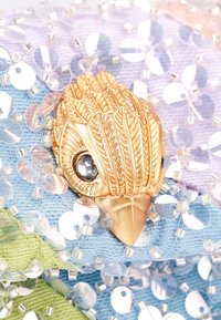 Decorazione dorata a forma di uccello con occhio di cristallo, circondata da paillettes iridescenti su tessuto pastello nelle tonalità di blu, verde e viola.