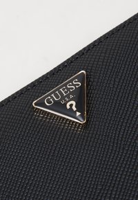 Svart texturerat material plånbok med en triangelformad logotyp med guldaccenter som visar "GUESS U.S.A." och en frågeteckendesign.