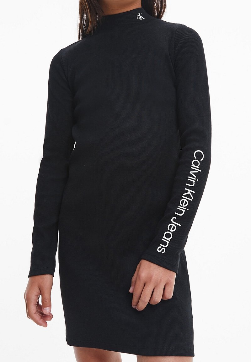 Abito lungo nero a maniche lunghe in tessuto a coste, con collo alto e testo "Calvin Klein Jeans" su una manica. Logo iconico sul colletto.