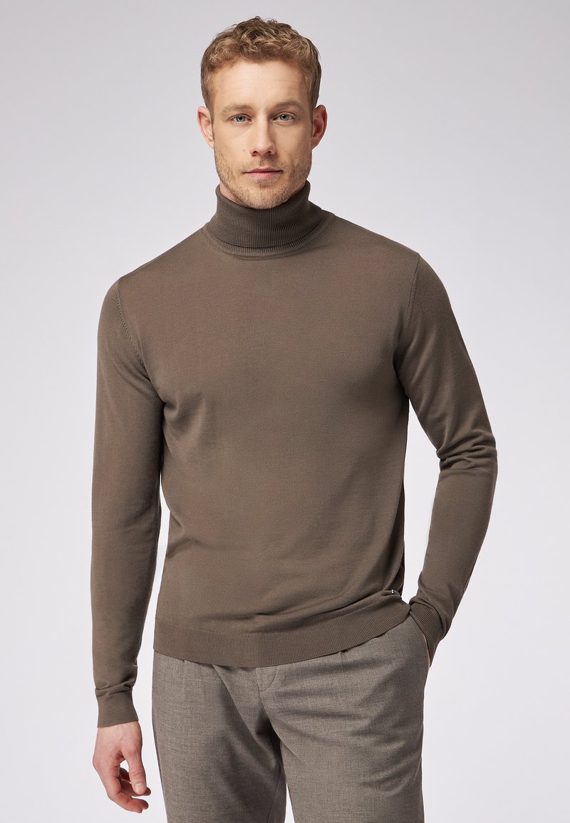 Maglione a collo alto marrone con texture a costine, maniche lunghe e design aderente. Abbinato a pantaloni grigi chiari.