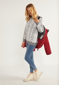 Lys grå sweater med snefnugsmønster, blå jeans og cremefarvede støvler. Rød puffjakke draperet over skulderen, der viser et afslappet outfit.