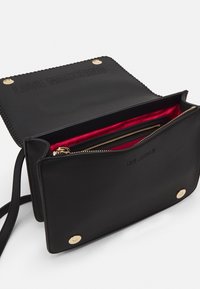 Černá kožená crossbody taška se zlatými kovovými prvky, texturovanými okraji a červeným textilním podšívkou. Obsahuje zipovou kapsu a embosované logo.