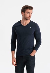 Uomo con barba che indossa una maglietta a maniche lunghe blu navy con scollatura a V e jeans scuri, in piedi con una mano in tasca, guardando di lato.