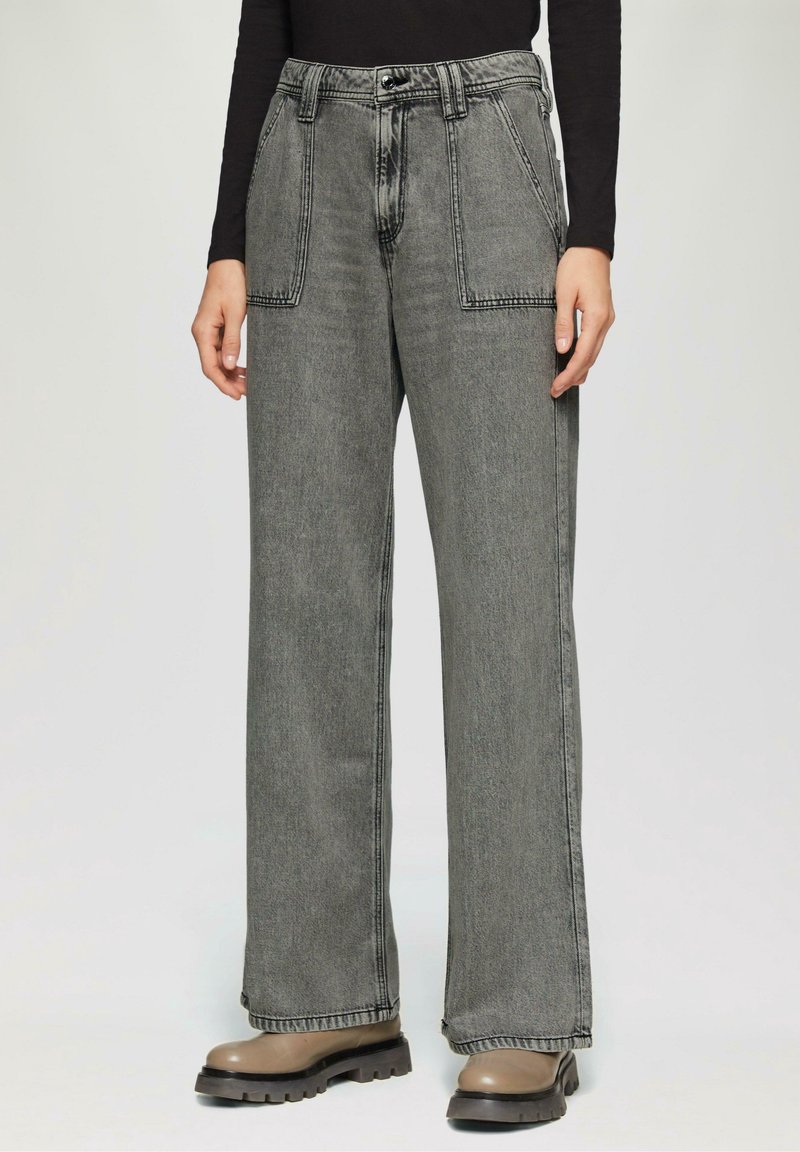 Personne portant un jean en denim gris taille haute et jambes larges avec de grandes poches plaquées à l'avant et des bottes beiges à semelle épaisse.
