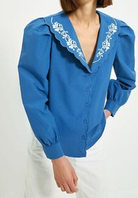 Blusa blu con bottoni, colletto sagomato, ricami floreali bianchi e maniche a sbuffo. Realizzata in tessuto morbido con una parte frontale aderente.