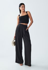 Sort crop top med tekstureret stof og justerbare stropper, parret med højtsiddende, viddebenede sorte bukser. Modellen holder en vævet taske.